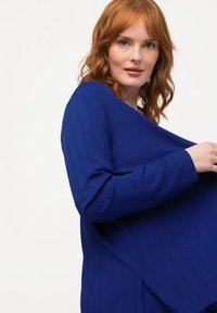 Blauw geribbeld long-sleeve top met een losse pasvorm en zijsplitten. Kenmerkt zich door een zachte textuur en een V-halslijn, weergegeven tegen een lichte achtergrond.