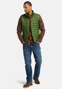 Grön vadderad väst med hög krage, bärs över en brun rutig skjorta. Blå jeans och bruna stövlar fulländar outfiten. Avslappnad, utomhusstil.