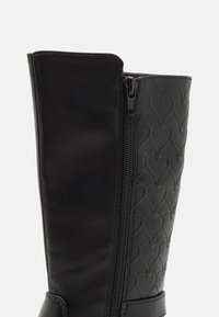 Bottines noires à hauteur de genou avec un tige en cuir lisse et un panneau en caoutchouc texturé avec des motifs en forme de cœur, accentuées par une fermeture éclair latérale.