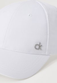 Hvit baseballcaps i bomull med glatt tekstur. Har en liten sølvfarget "ck"-logo på siden og ventilasjonshull.