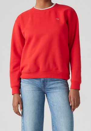 Persoon draagt een felrood crewneck sweatshirt met een klein logo op de borst en lichtblauwe spijkerbroek tegen een effen achtergrond.