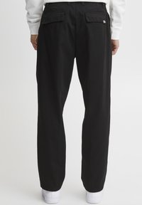 Pantaloni negri cu croi drept, având două buzunare spate și un etichetă logo subtilă; asortați cu adidași albi, pe un fundal simplu.