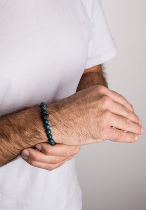 Beaded bracelet featuring round teal and black stones, with a textured surface, worn on a wrist against a plain white t-shirt.