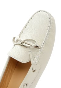 Mocasín de cuero blanco con superficie texturizada, herrajes plateados y un acento de lazo. Presenta costuras en contraste y un diseño abierto.