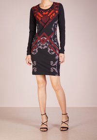 Robe noire à manches longues avec des motifs rouges et gris complexes, col rond et silhouette ajustée, accompagnée de talons noirs à lanières.