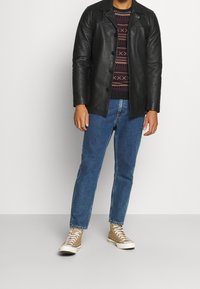 Veste en cuir noire à fermeture par boutons, pull à motifs, jean en denim bleu et baskets beiges à lacets. Tenue décontractée en superposition.