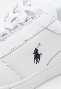 Vit lädersneaker med texturerad yta, rund tå och svart broderad logotyp. Har snörning och vit gummisul.