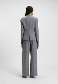Donna con lunghi capelli intrecciati che indossa un blazer grigio su misura e pantaloni a gamba larga, in piedi di spalle alla fotocamera su uno sfondo neutro.