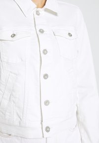 Chaqueta de mezclilla blanca con un diseño corto, que presenta botones a presión plateados, dos bolsillos en el pecho y un cuello puntiagudo. Detalle de tela texturizada visible.