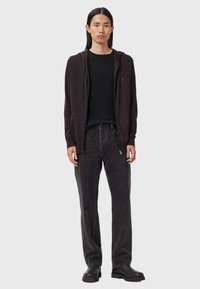 Ung man med långt hår som bär en mörk zip-up hoodie, svart t-shirt, urblekta svarta jeans och svarta stövlar, står mot en enkel bakgrund.