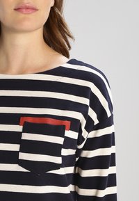 Camisetta a righe blu navy e bianche con collo rotondo e taschino sul petto con un accento rosso. Realizzata in tessuto morbido e testurizzato.
