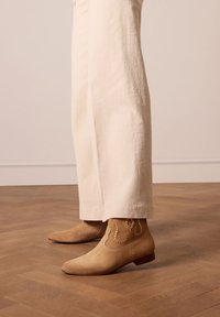 Café suède enkellaarsjes met puntige neuzen, open stiksel detail en een lage hak, gedragen met crème wijde broek op een houten vloer.