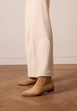 Braune Wildleder-Ankle Boots mit spitz zulaufenden Zehen, offenen Nahtdetails und einem niedrigen Absatz, getragen zu cremefarbenen, weit geschnittenen Hosen auf einem Holzboden.