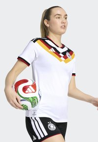 White soccer jersey with red, black, and yellow accents, featuring a V-neck, short sleeves, and a logo. Black shorts with white stripes. Multi-colored ball.