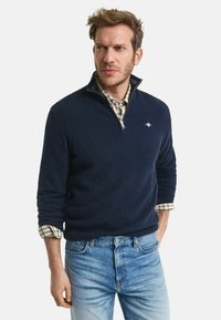 Navy Zip-Up-Pullover aus strukturiertem Strickstoff, mit Kragen und kleinem Logo, über einem karierten Hemd mit hochgerollten Ärmeln getragen.