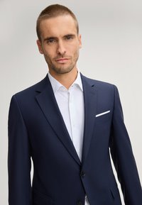 Abito blu navy con rever a incavo; camicia da uomo bianca con pois; taschino al petto singolo; bottoni neri; tessuto dalla trama liscia.