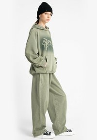Młoda kobieta ubrana w luźny, wyblakły zielony hoodie i dopasowane szerokie spodnie, w czapce beanie w kolorze czarnym oraz czarno-białych sneakersach, stojąca bokiem.