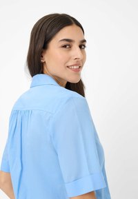Camicia azzurra a maniche corte con colletto e dettaglio di piega arricciata sul retro. Tessuto liscio con una consistenza morbida.