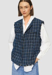 Blauer und schwarzer Tweed-Weste mit Fransenbesatz, ausgestattet mit zwei Fronttaschen und einem Knopfverschluss, getragen über einem weißen Hemd.