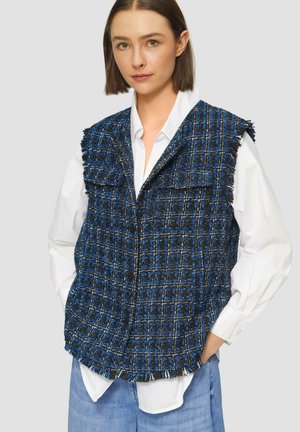 Blauwe en zwarte tweed gilet met franjes, voorzien van twee voorzakken en een knoopsluiting, gedragen over een wit shirt.