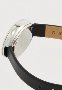 Ασημένια μεταλλική κάσα ρολογιού με μαύρη δερμάτινη λωρίδα και μπεζ κάτω μέρος. Περιλαμβάνει κορώνα και χαραγμένες λεπτομέρειες στη πίσω πλευρά της κάσας.