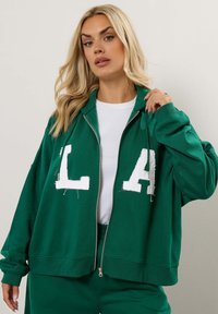 Felpa verde con zip e scritta bianca "LA", dal taglio ampio e rilassato. Realizzata in tessuto morbido con polsini a coste e zipper frontale.