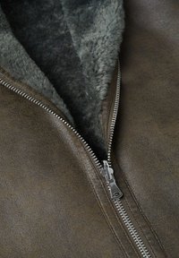 Bruine nepleer jas met een grijze bontomzoomde kraag en zilveren rits, met een gestructureerde buitenkant en een gladde afwerking.