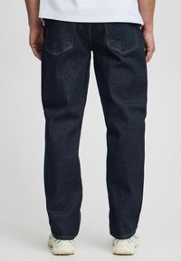 Dark denim jeans with a straight cut, featuring two back pockets and visible stitching. Worn with a white shirt and light sneakers.