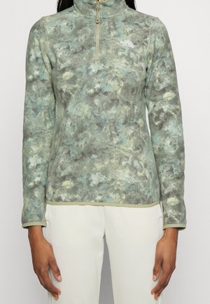 Grün-grauer marmorierter Fleece-Pullover mit Viertelreißverschluss und weißem Logo, getragen zu weißen Hosen.