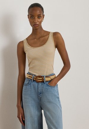 Naine, kes kannab beeži ja valge triibulist varrukateta pluusi, kõrge vöökohaga helerohelisi teksapükse ning punutud beeži vööd puidust pandlaga.