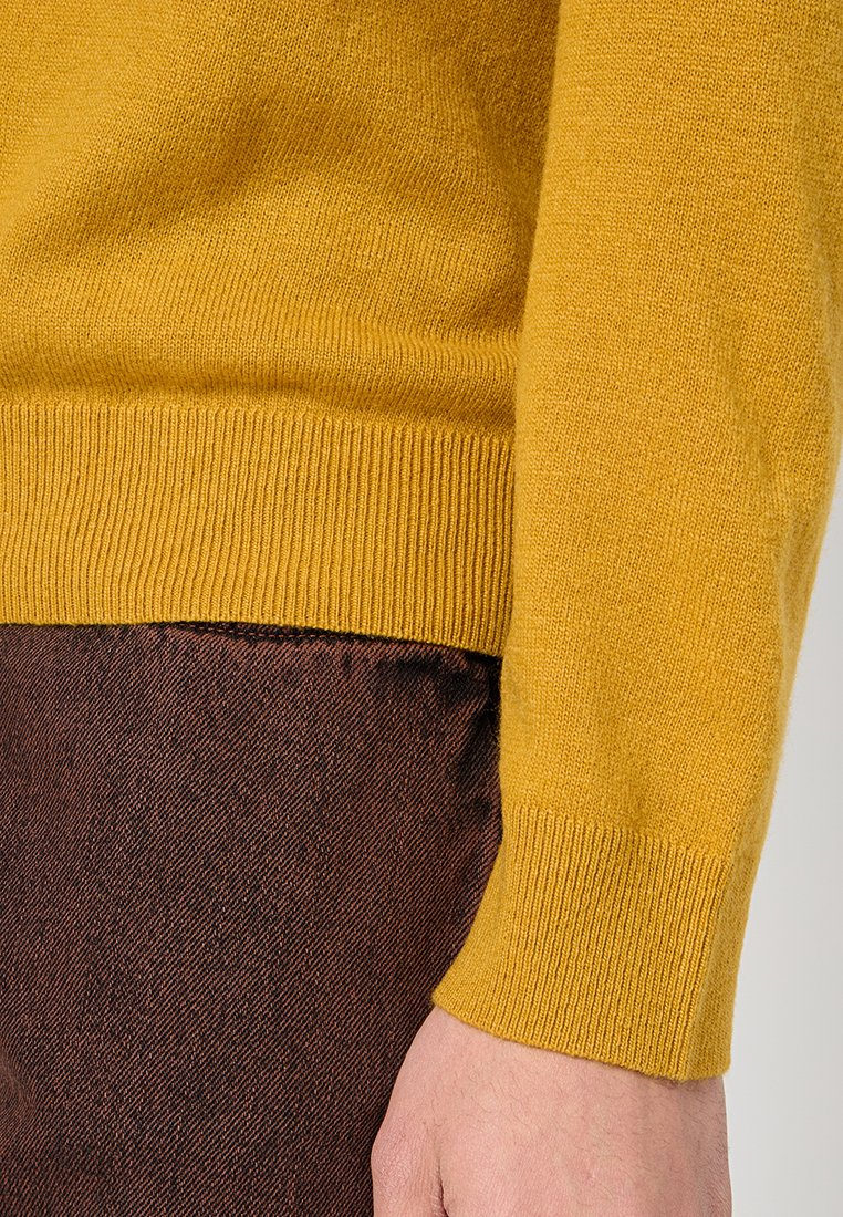 Gul stickad tröja med ribbade ärmar, visad nära bruna texturerade byxor. Tröjan har ett mjukt, medeltjockt material.