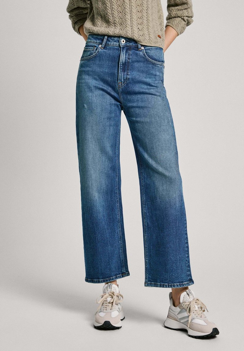 Laiad sinised denim püksid keskmise kõrgusega talje ja minimaalsete kulumismärkidega, koos heledate spordijalatsitega. Ülalpool on struktuurne keeratud kampsun.