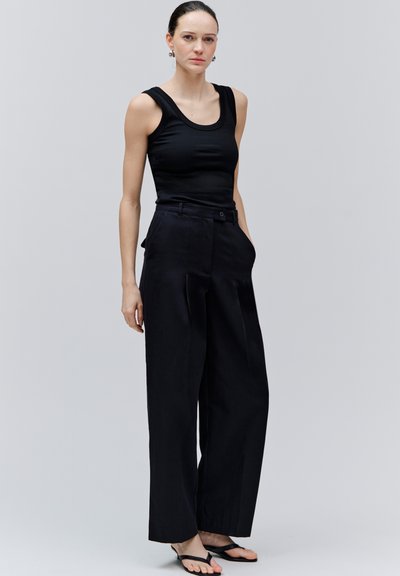 Vrouw met een zwarte, mouwloze top, zwarte wijde broek en zwarte sandalen, staand tegen een effen lichte achtergrond.