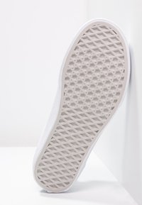 Semelle de sneaker blanche avec un motif de diamonds et d'étoiles, inclinée contre un mur et une surface blanche.