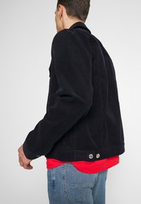 Chaqueta de denim de corduroy negra con un corte relajado, que presenta dos botones de metal en la cintura y una textura ribeteada visible. Lleva puesta una camisa roja.