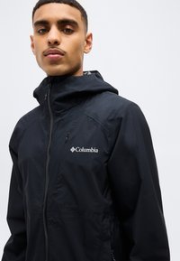 Άνδρας που φοράει μαύρο μπουφάν Columbia με κουκούλα, φερμένο μέχρι πάνω, κοιτάει μπροστά με φόντο λείο, ανοιχτόχρωμο τοίχο.