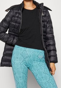 Svart pufferjacka med blank finish och dragkedja, kombinerad med en figurnära svart topp och turkosa leggings med ormmönster.