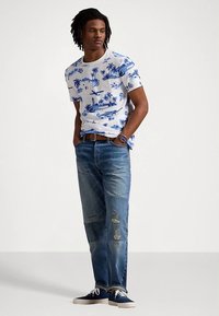 Λευκό μπλουζάκι με μπλε τροπικό σχέδιο, συνδυασμένο με ανοιχτό μπλε τζιν με φθορές. Το μοντέλο φοράει σκούρα αθλητικά παπούτσια και καφέ ζώνη.