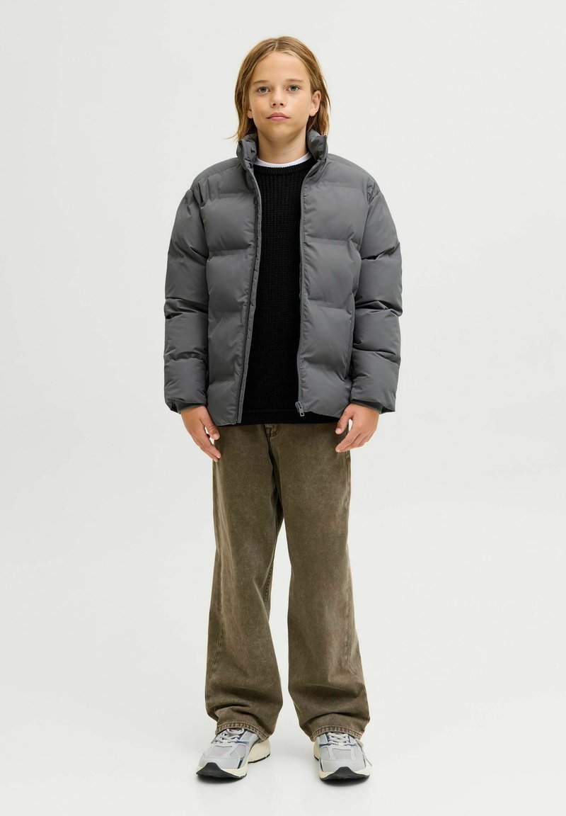 Grå polstret jakke med høj krave, båret over en sort sweater. Kombineret med olivengrønne bukser og grå sneakers. Neutral baggrund.