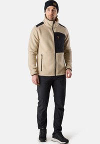 Beige fleece jacket with black accents and zippered chest pocket, paired with black pants and black sneakers, presented against a white background.