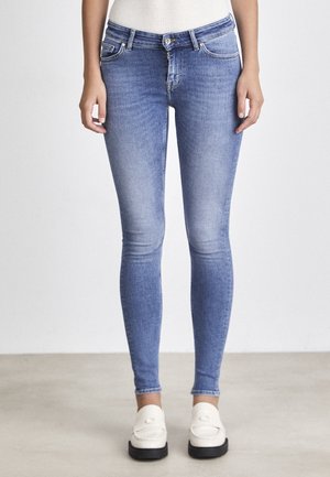 Slim-fit blå jeans båret med hvide loafers og en hvid ribbet top, vist fra taljen til fødderne mod en ensfarvet baggrund.