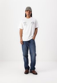 White cotton t-shirt with printed text, paired with blue jeans and brown slip-on shoes. A gray cap with embroidered text completes the outfit.