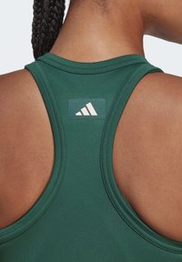 Canotta sportiva verde con scollatura rotonda e design a schiena scoperta. Presenta un pannello con il logo Adidas beige sulla parte superiore della schiena. Tessuto di trama liscia.