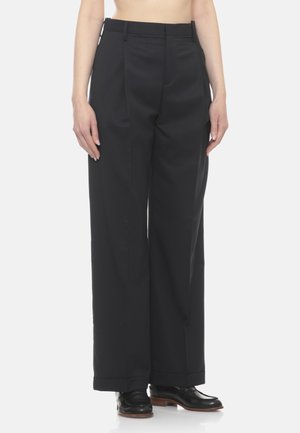 Pantaloni neri a vita alta e gamba larga indossati da una persona, abbinati a scarpe di pelle nere con suole marroni, mostrati dalla vita ai piedi.