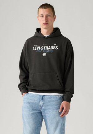 Młody mężczyzna o krótkich włosach, ubrany w czarną bluzę z kapturem Levi Strauss i jasnoniebieskie dżinsy, stojący na jednolitym jasnym tle.