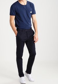 Mężczyzna w granatowej koszulce, ciemnoniebieskich slim-fit spodniach w białe kropeczki i białych trampkach, stojący z jedną ręką w kieszeni.