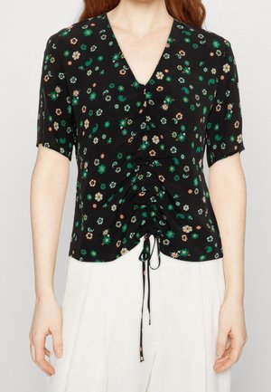 Blusa nera con fiori verdi e bianchi, maniche corte, scollatura a V e coulisse arricciata davanti, abbinata a pantaloni bianchi a vita alta.