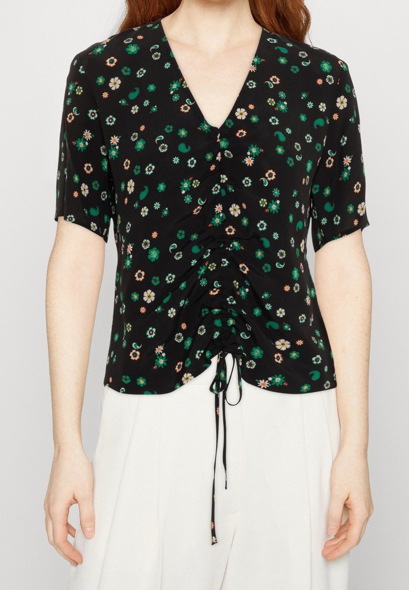 Blusa nera con fiori verdi e bianchi, maniche corte, scollatura a V e coulisse arricciata davanti, abbinata a pantaloni bianchi a vita alta.