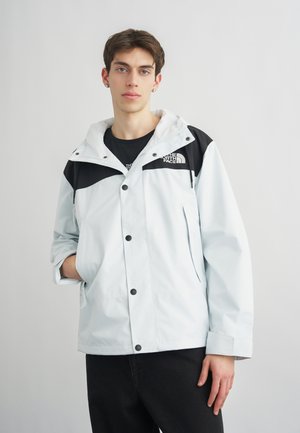 Νέος άνδρας που φοράει ένα ανοιχτό γκρι και μαύρο μπουφάν North Face με κουκούλα, στέκεται μπροστά σε ένα απλό ανοιχτό γκρι φόντο.