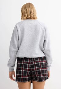 Sudadera gris con mangas raglán y escote redondeado, combinada con pantalones cortos negros con un patrón de cuadros en rojo y blanco. Fondo liso.